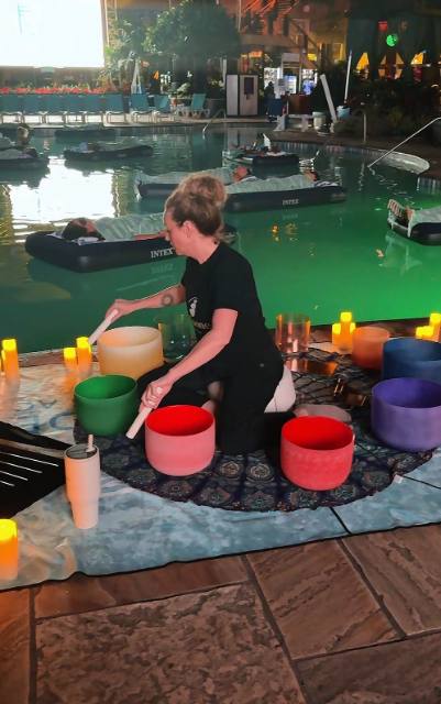 Healer playing crystal singing bowls poolside while guests float on rafts
